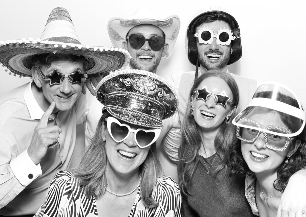 large group of friends using the photo booth with email collection feature, during a brand launch, while having fun and posing with props and hat, with a b&w filter on