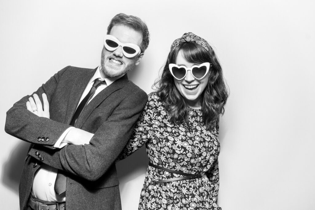 black and white image of bride and groom posing holding eachother wearing hats with a white backdrop during a wedding party entertainment for mad hat photo booth rental in the cotswolds