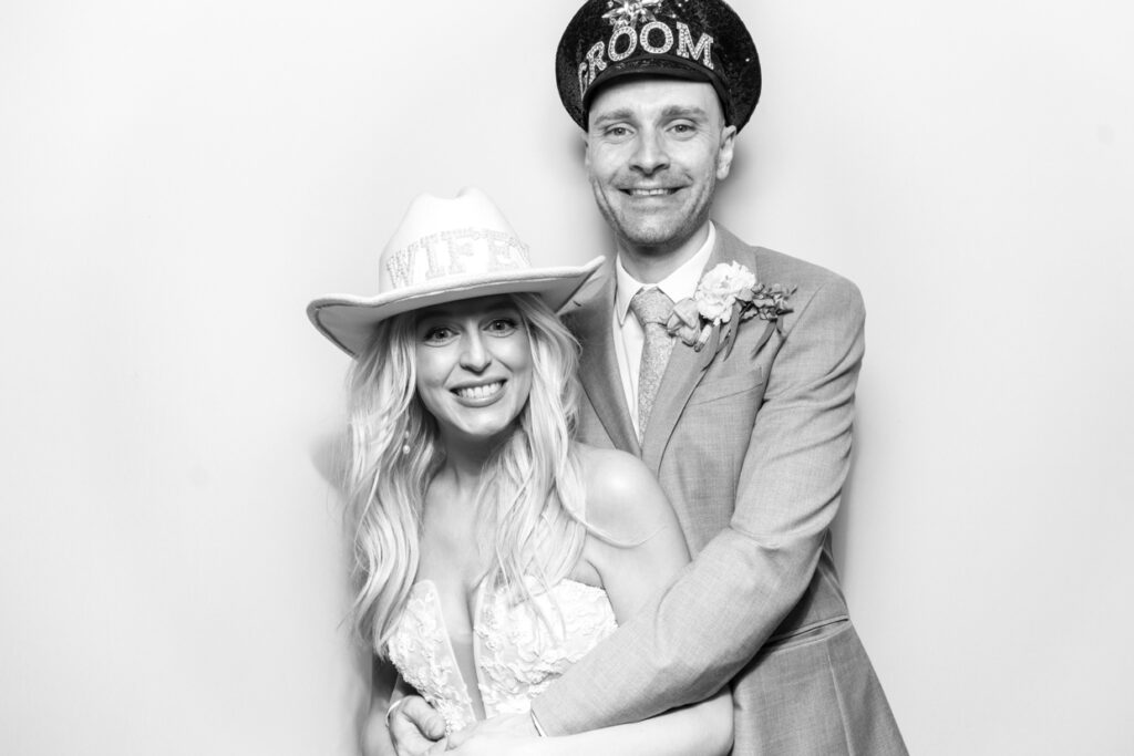 black and white image of bride and groom posing holding eachother wearing hats with a white backdrop during a wedding party entertainment for mad hat photo booth rental in the cotswolds