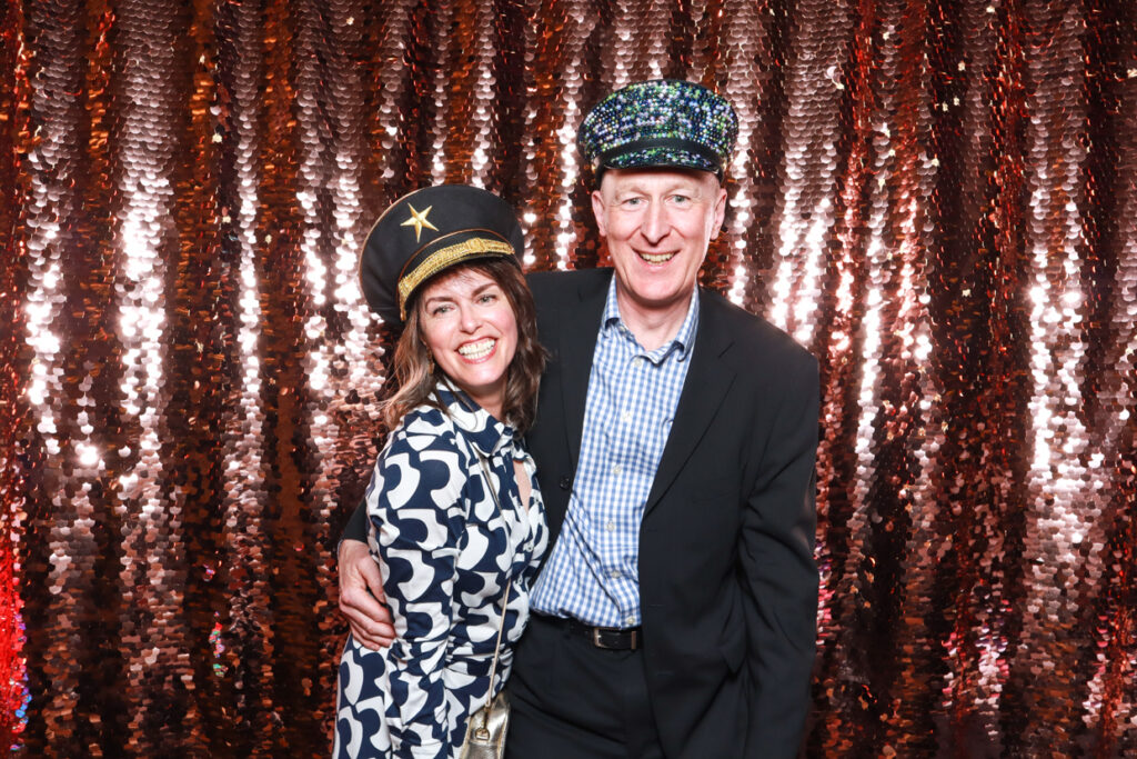 2 guests wearing fun props and hats posing against a champagne sequins backdrop during a brand event for a photo booth party rental 