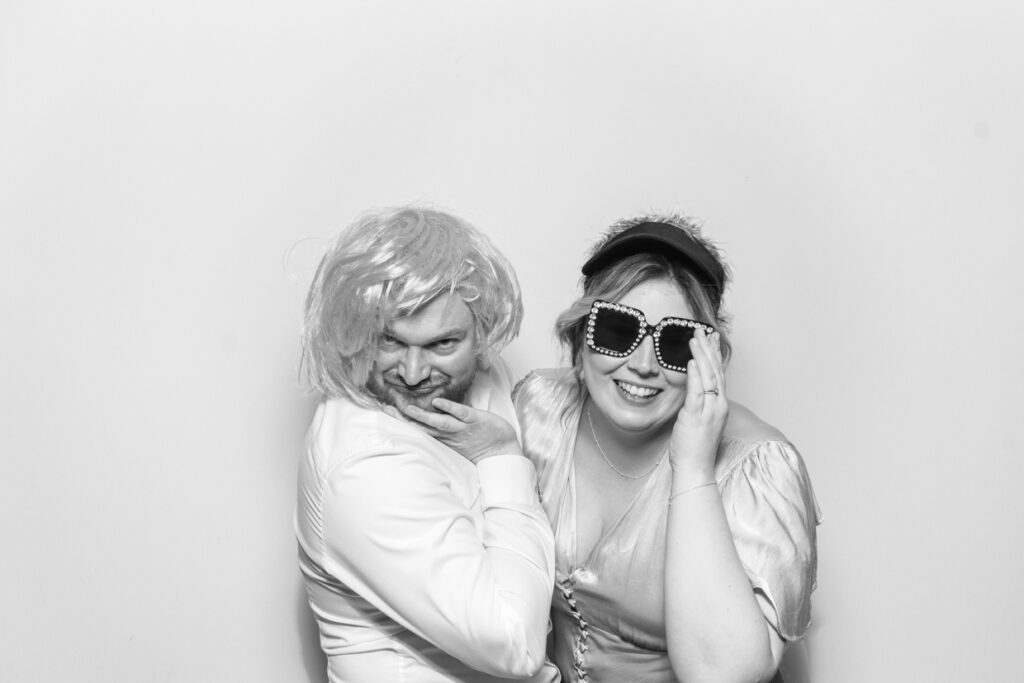 black and white image for couple posing in a funny way wearing props and wigs with a white backdrop during a wedding party entertainment for mad hat photo booth rental in the cotswolds