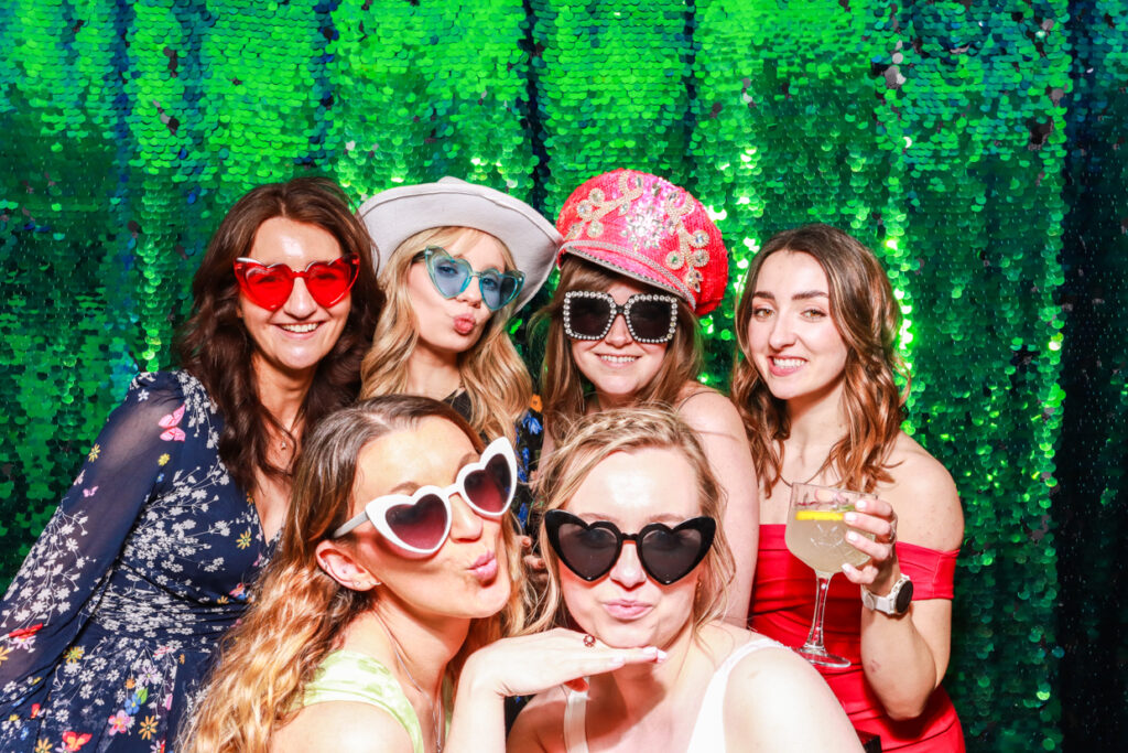 large group of friends including the bride wearing fun props and hats posing against a green sequins backdrop during a wedding reception party entertainment for a photo booth party rental 