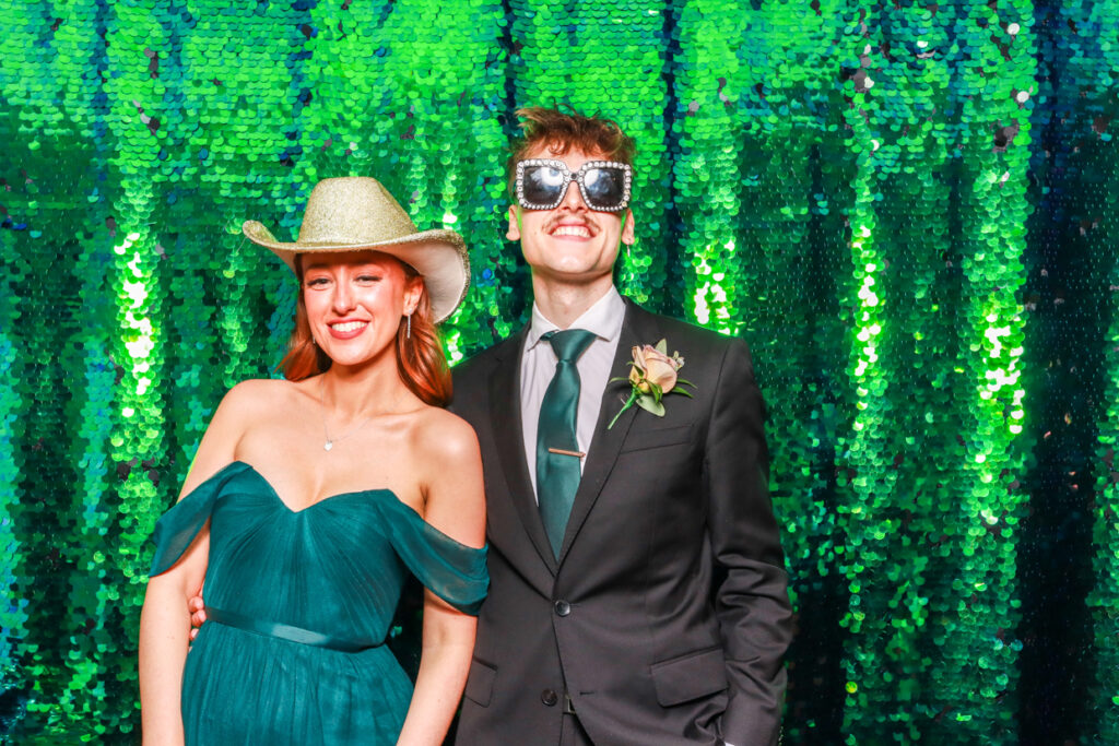 couple wearing fun props and hats posing against a green sequins backdrop during a corporate party entertainment for a photo booth party rental 