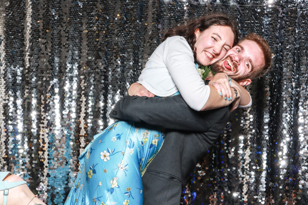 boy hugging and lifting up girl while posing in a fun way for mad hat photo booth, with a silver backdrop behind during a private event entertainment hire