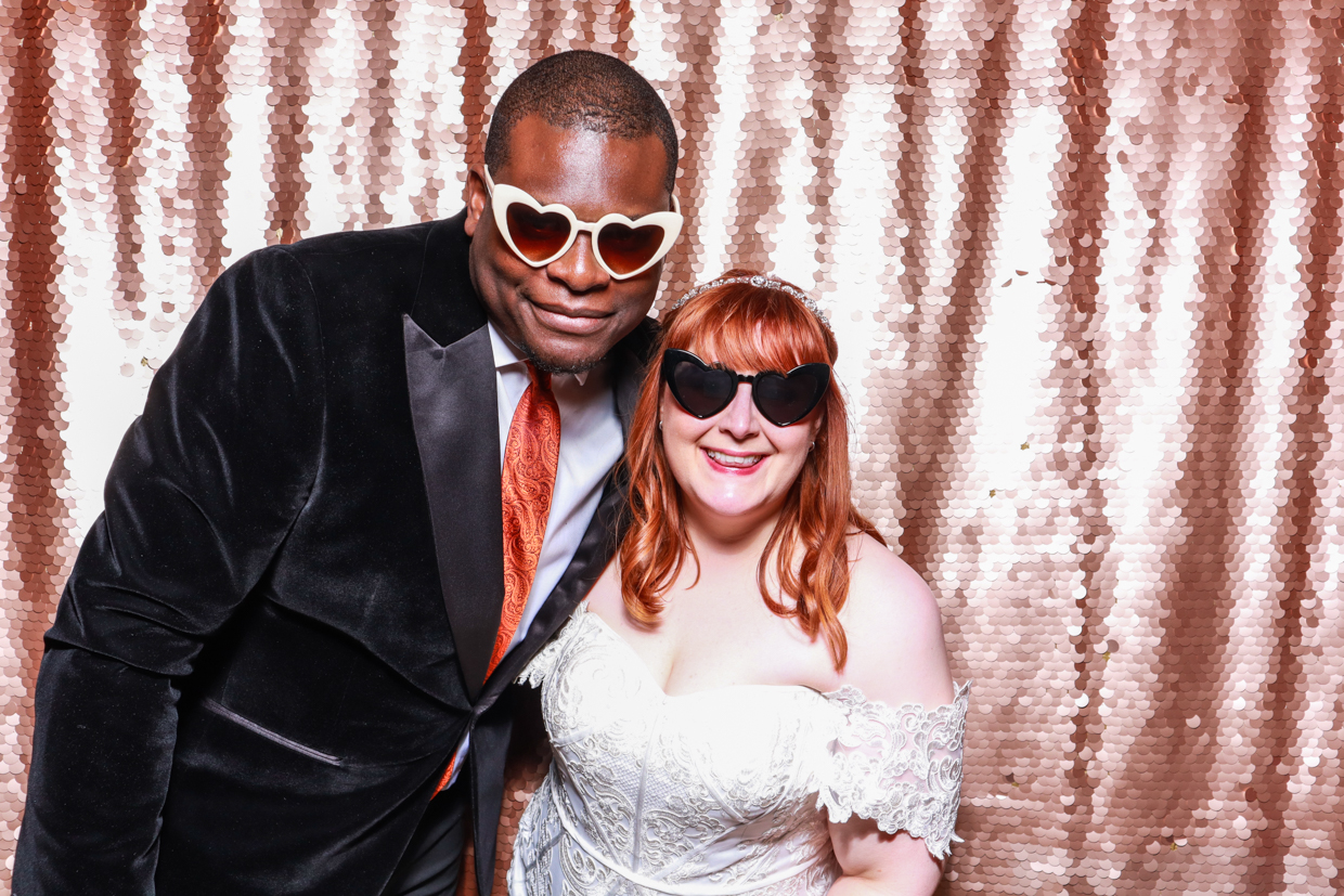 bride and groom wearing a velvet suit and a bridal dress, posing against a blush sequins shiny backdrop during a wedding party, wearing fun heart shaped sunglasses 