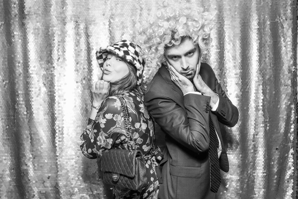 b&w print of 2 guests wearing fun props and posing against a silver sequins background, during a private party event with an Affordable Christmas Photo Booth Rental 