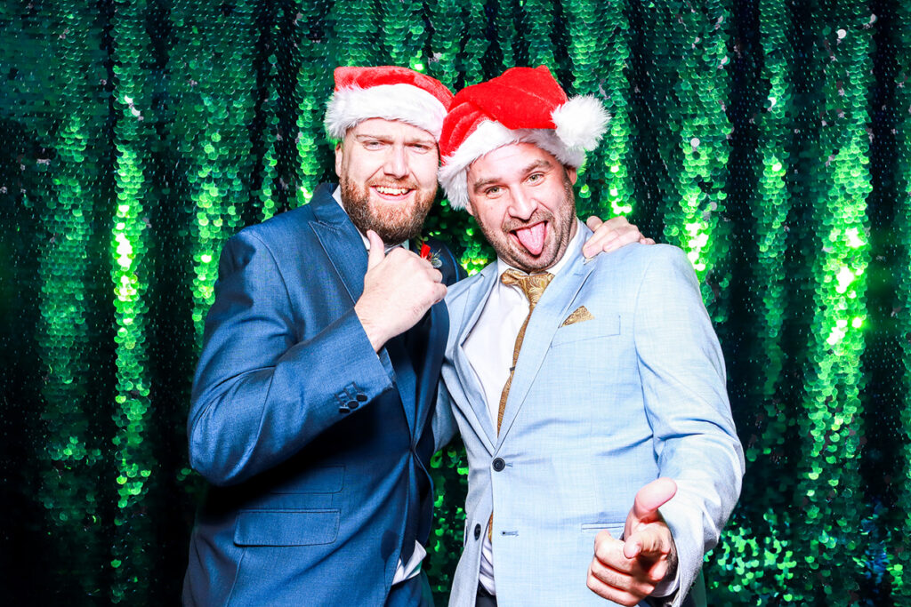 2 guests posing against a green sequins backdrop, wearing Santa hats during a corporate staff party 