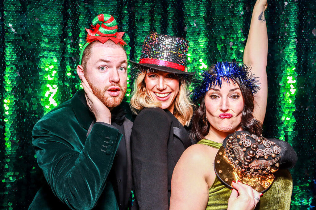 3 guests posing against a green sequins backdrop, wearing Santa hats and festive props, during a corporate staff party with our mad hat photo booth rental