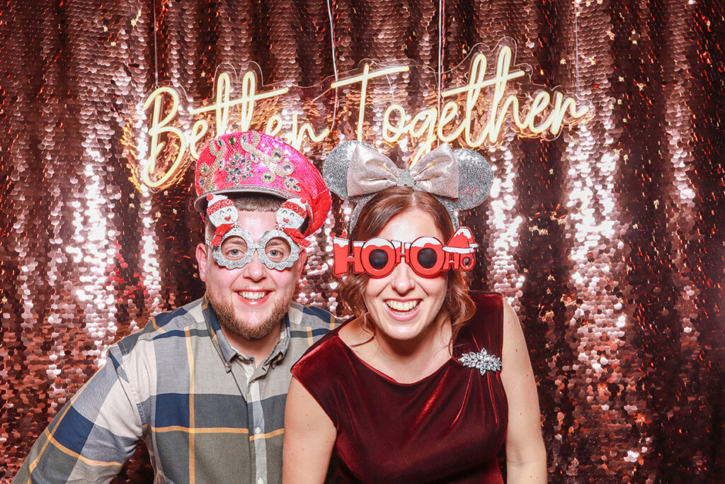 2 guests posing against a champagne sequins backdrop, wearing Santa hats during a corporate staff party during Christmas