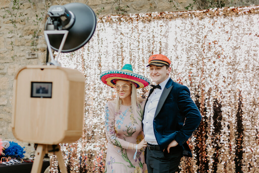 2 guests during a party entertainment with mad hat photo booth, posing against a champagne sequins backdrop for a corporate event in chipping campden, Cotswolds