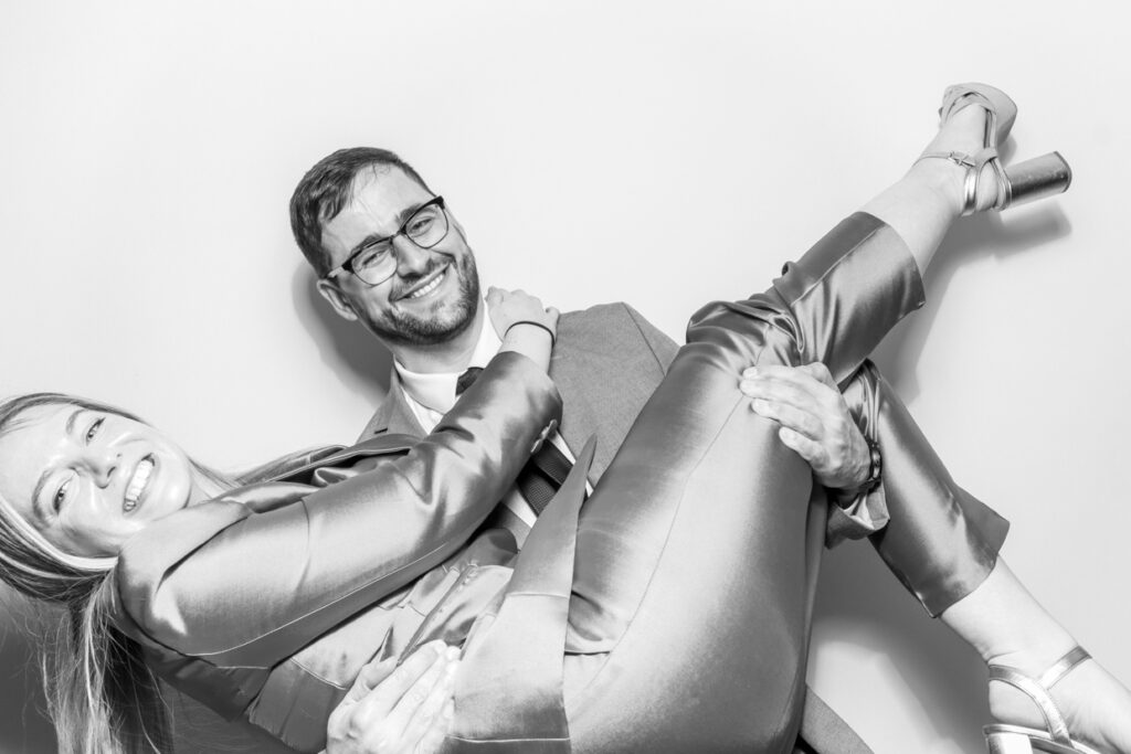 black and white photo 2 guests lifting the other up, during a corporate event party entertainment, with the couple posing against a white backdrop, for a kardashian style glam unlimited print offer by mad hat photo booth