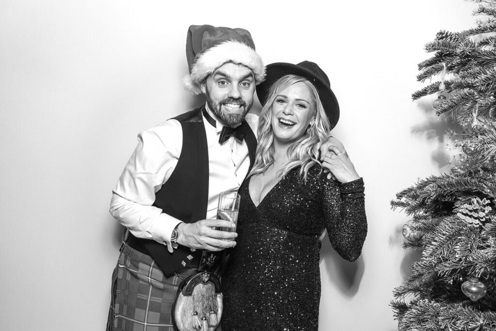 black and white print of 2 guests against a white backdrop, posing near a christmas tree, during a party entertainment event in the cotswolds