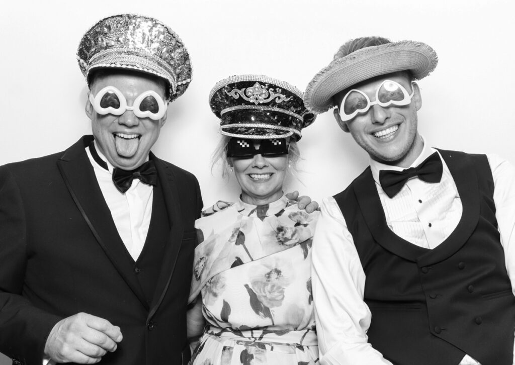 3 guests posing against a white backdrop with mad hat photo booth, using fun props during a corporate event party entertainment 