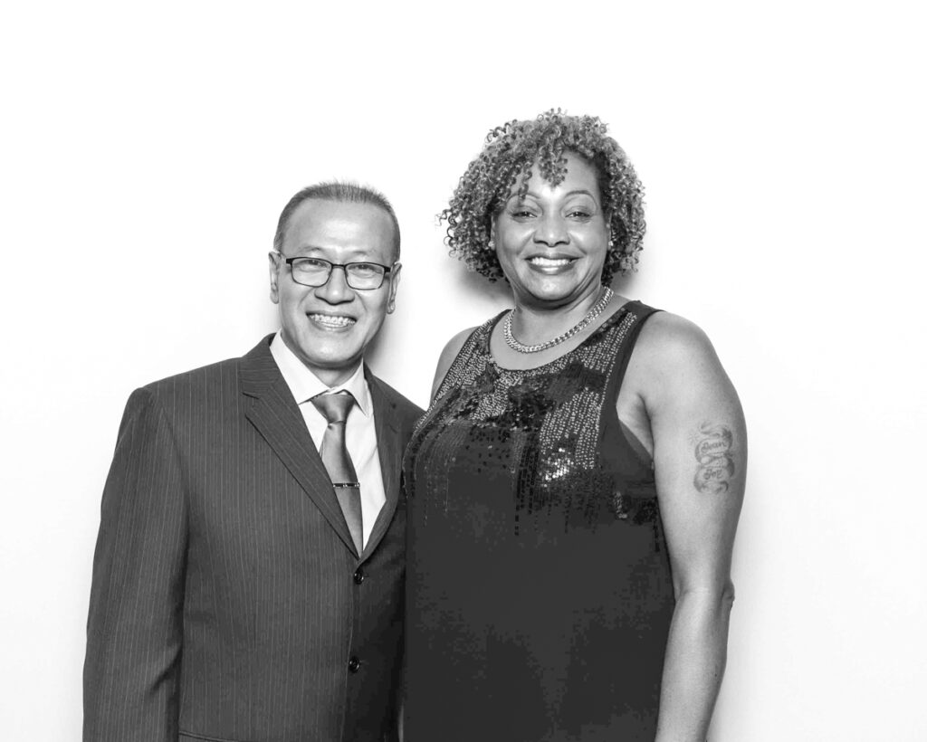 Black and white photo of a man in a suit and tie standing next to a woman in a sequined top, both smiling warmly at a Cheltenham Racecourse event