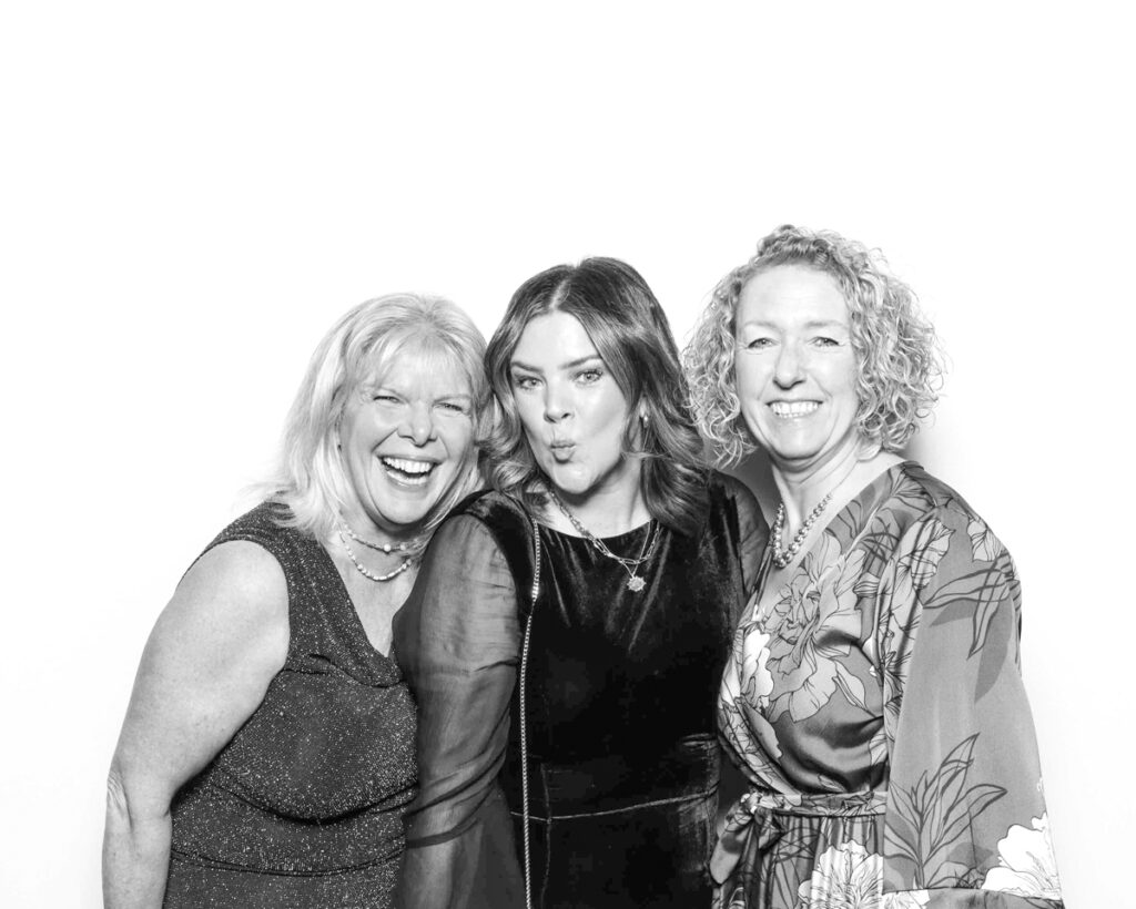 Black and white group photo of three women standing close together, smiling warmly, dressed in elegant formal wear, during a corporate event photo booth party entertainment