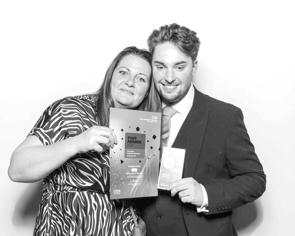 Black and white group photo of 2 people holding a NHS Staff Awards 2024 diploma, standing close together, smiling warmly, during a corporate event in Gloucestershire