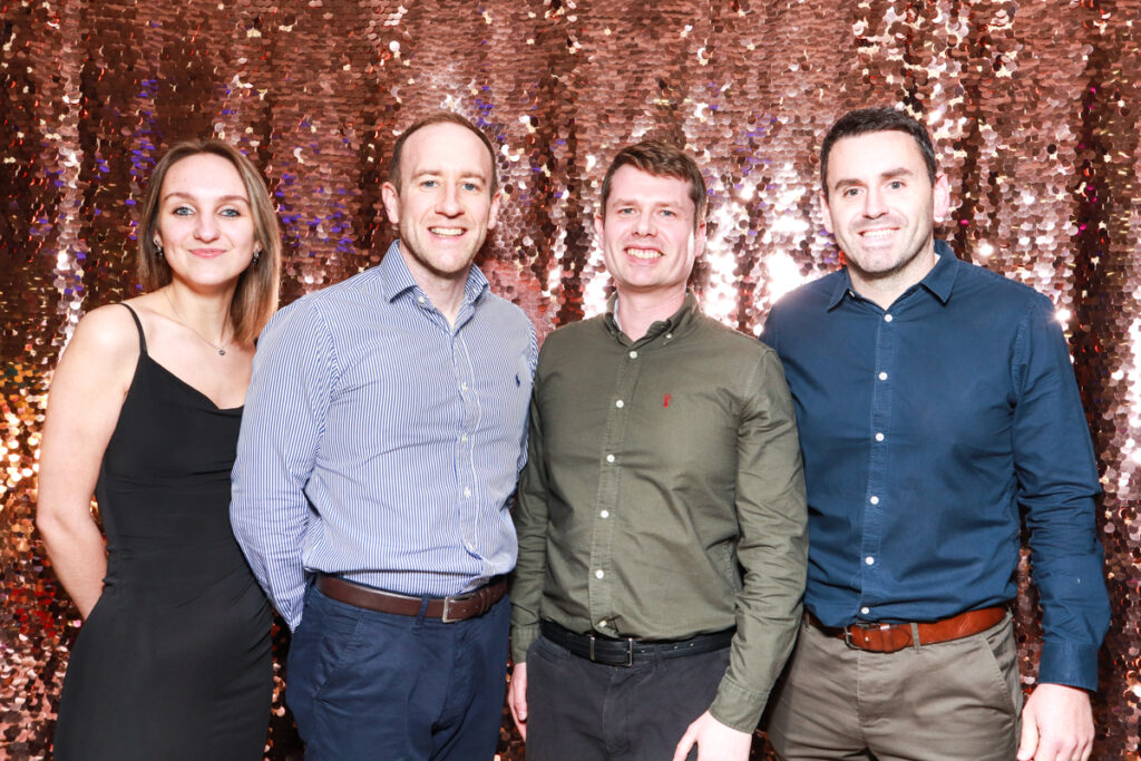 Group photo of three men and a woman standing in front of a champagne sequins backdrop, dressed smart-casual and smiling warmly at a corporate event at Cheltenham Racecourse.