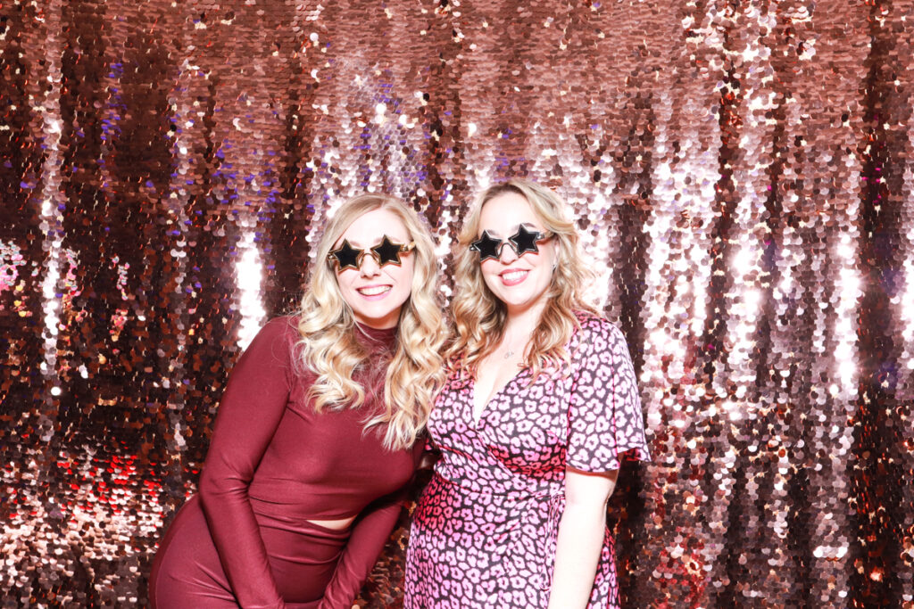 Two women wearing star-shaped glasses and smiling playfully in front of a champagne sequins backdrop at a Cheltenham Racecourse event.