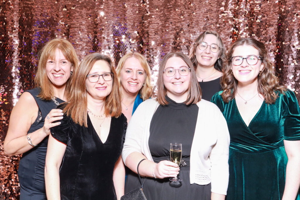 Group photo of six women dressed in evening wear, smiling and holding glasses of champagne in front of a champagne sequins backdrop at a Cheltenham Racecourse corporate event.