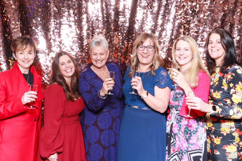 Group photo of six women dressed in colourful formal outfits, smiling and holding drinks in front of a champagne sequins backdrop at a corporate event at Cheltenham Racecourse."