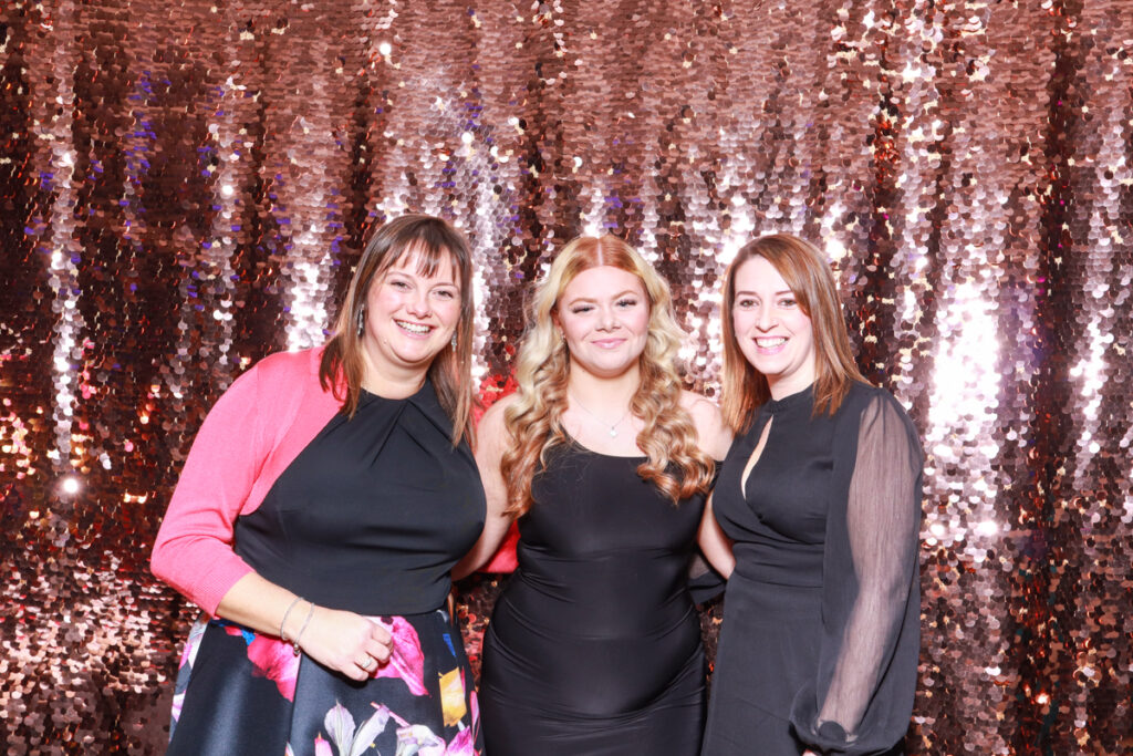 Three women dressed in elegant black and colourful evening wear, smiling in front of a champagne sequins backdrop for a corporate event staff party at Christmas