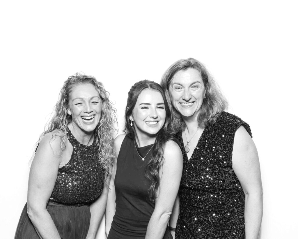 Black and white photo of three women in sparkling dresses, smiling and laughing in front of a plain white backdrop at a Cheltenham Racecourse event."