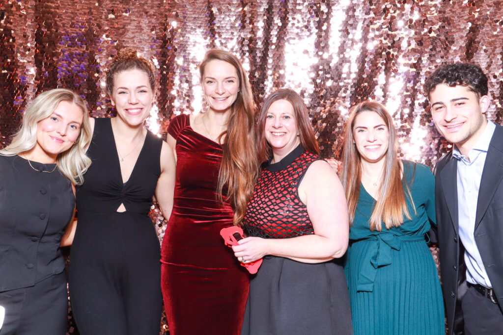 Group photo of six colleagues dressed elegantly, smiling in front of a champagne sequins backdrop at a Cheltenham Racecourse corporate event.