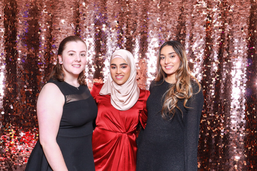 Three women in stylish outfits, including a red dress with a hijab, smiling in front of a champagne sequins backdrop for a Christmas Staff party entertainment