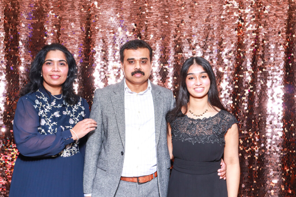 Family group photo featuring two women and one man in formal attire, smiling in front of a champagne sequins backdrop at a corporate event at Cheltenham Racecourse