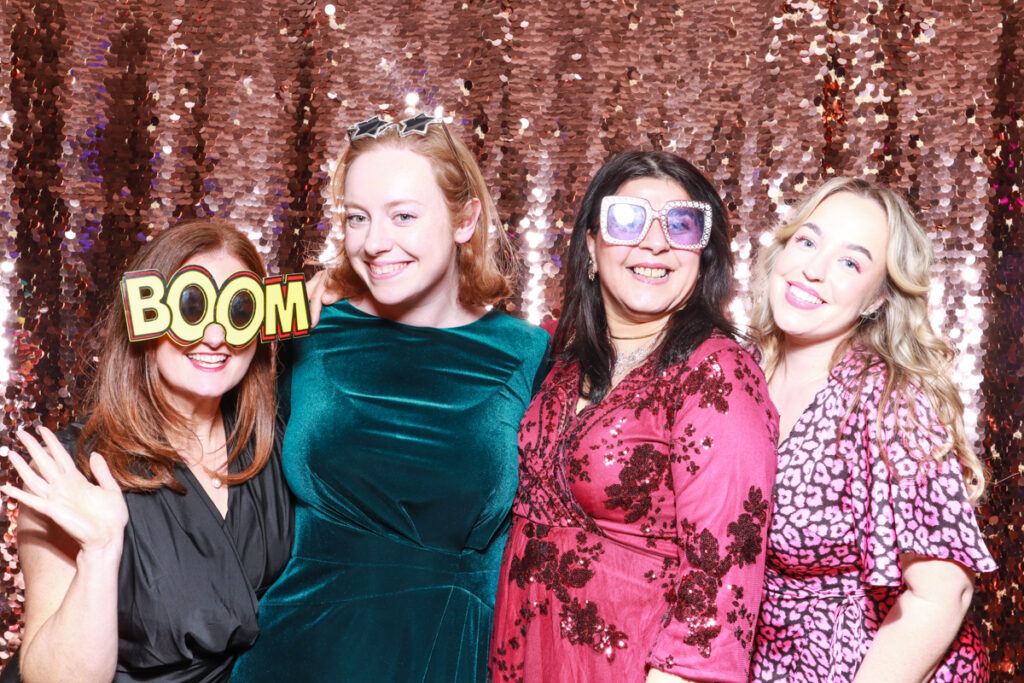 Four women dressed in bright and vibrant outfits, playfully posing with novelty glasses and props in front of a champagne sequins backdrop at a Cheltenham Racecourse event."