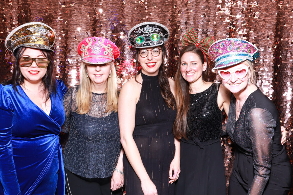 Five women wearing sparkling hats and glamorous attire, posing and smiling in front of a champagne sequins backdrop for a corporate party entertainment during Christmas