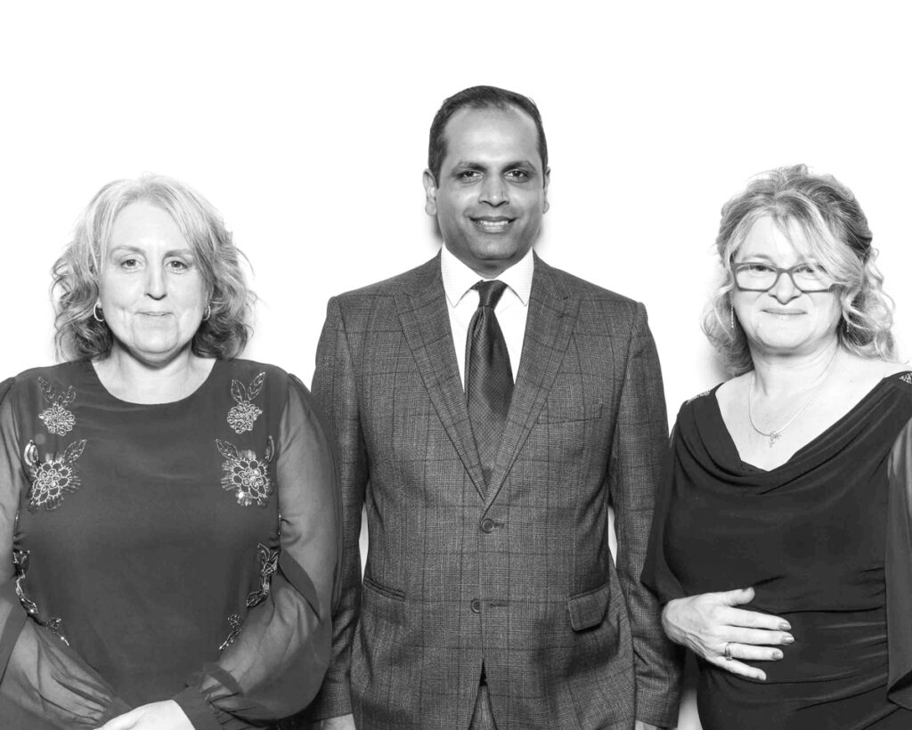 Black and white photo of a man in a suit standing between two women in formal dresses, all smiling at a Cheltenham Racecourse corporate event."