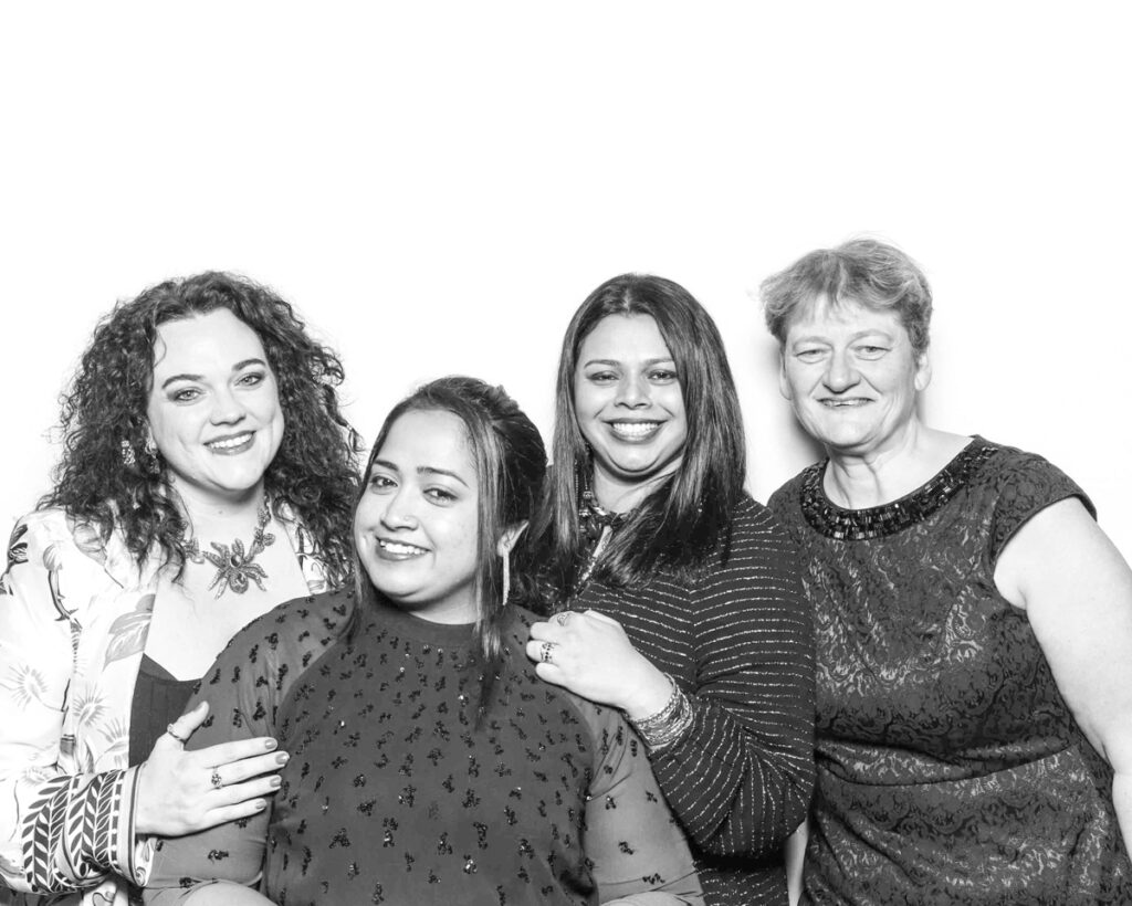 Black and white group photo of four women smiling, wearing a mix of formal and patterned outfits, captured at a Cheltenham Racecourse event."