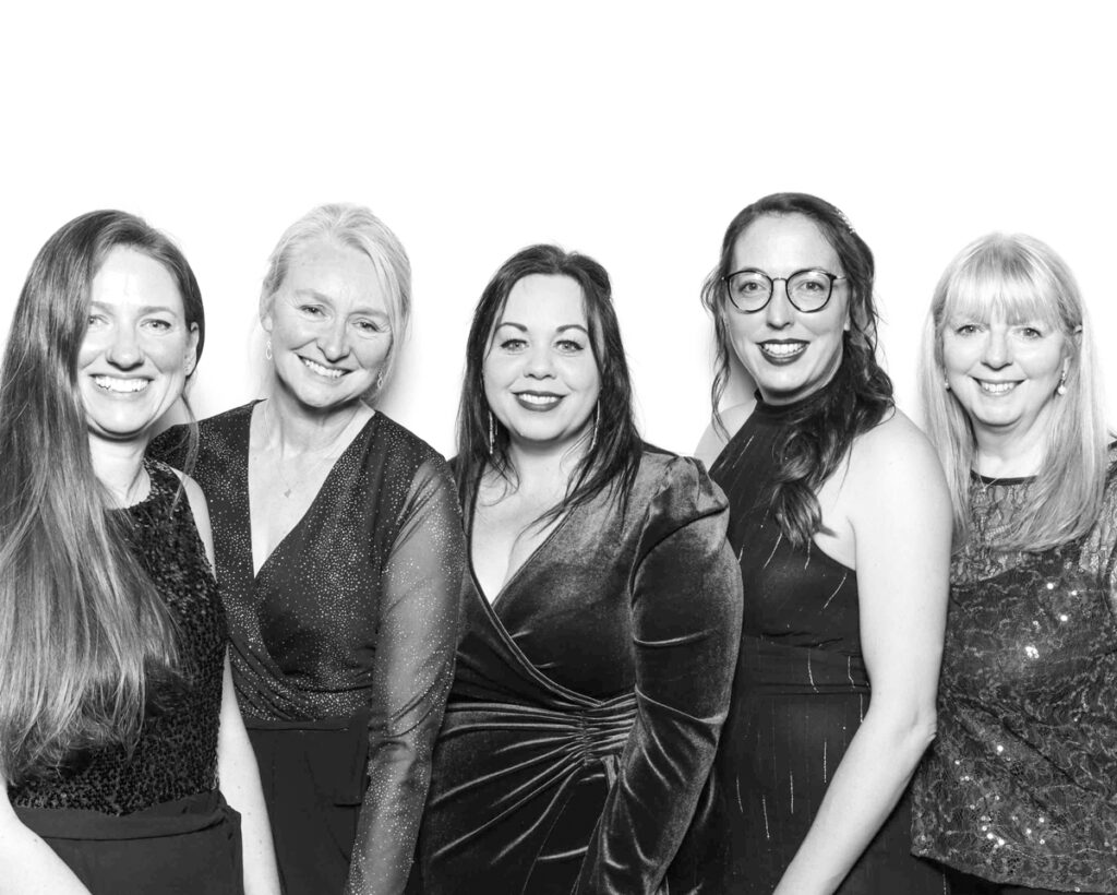 Black and white group photo of five women dressed elegantly in evening attire, smiling for the camera against a plain white backdrop. Captured at a corporate event at Cheltenham Racecourse with Mad Hat Photo Booth attending the event 