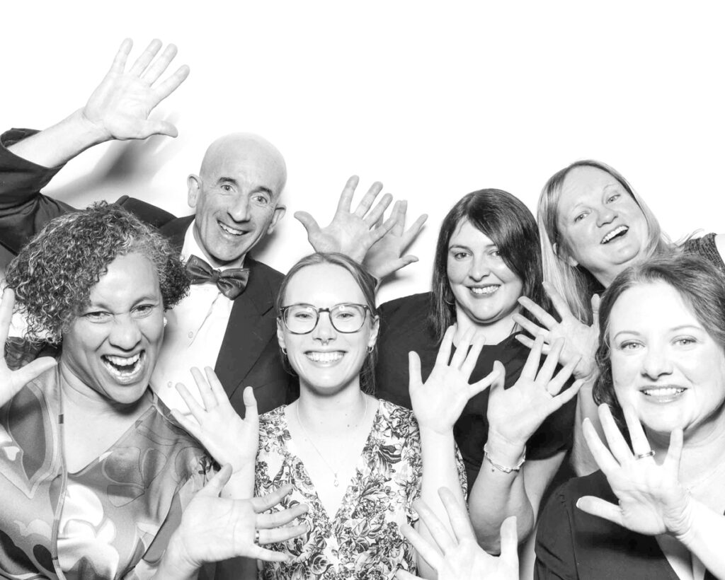 Black and white group photo of six people dressed in formal attire, smiling and laughing with their hands up in the air, in front of a plain white backdrop at a Cheltenham Racecourse event."