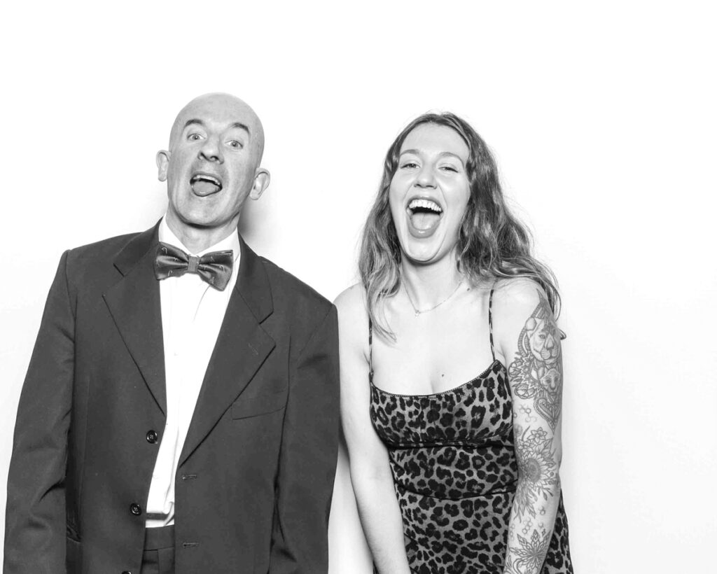 Black and white photo of a man in a tuxedo and a woman in a leopard print dress, both laughing playfully in front of a plain white backdrop at a Cheltenham Racecourse event.