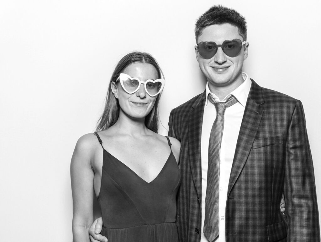 A stylish couple wearing heart-shaped sunglasses, posing in a sleek black and white photo booth, highlighting modern corporate event trends.