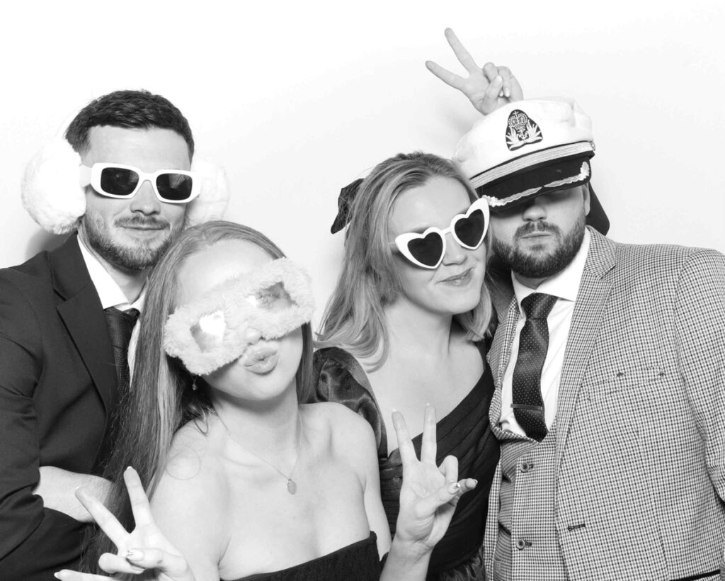 A group of four guests dressed formally, accessorised with quirky hats and playful sunglasses, striking fun poses in a black and white photo booth for a corporate event.