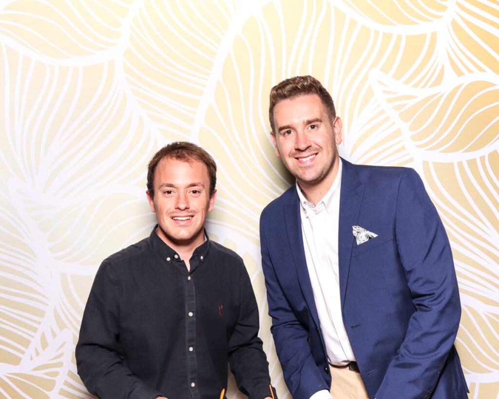 Two men dressed smartly, posing in front of a stylish gold backdrop, showcasing modern photo booth setups for corporate event trends.
