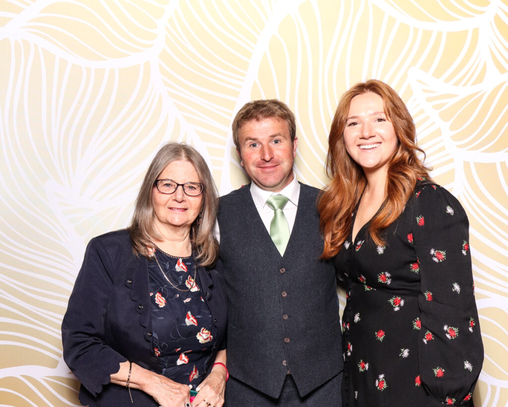 A group of three smiling guests, dressed formally, posing against a gold-patterned photo booth backdrop