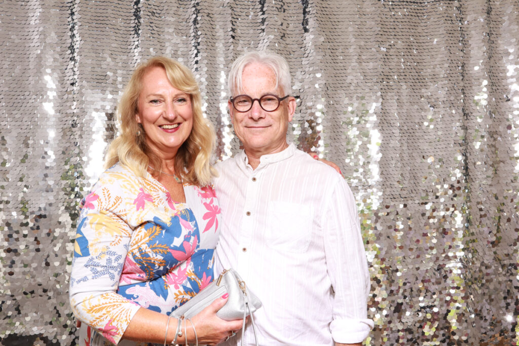 A couple dressed in vibrant outfits, posing against a glittering silver sequin backdrop, highlighting creative corporate event photo booth trends.