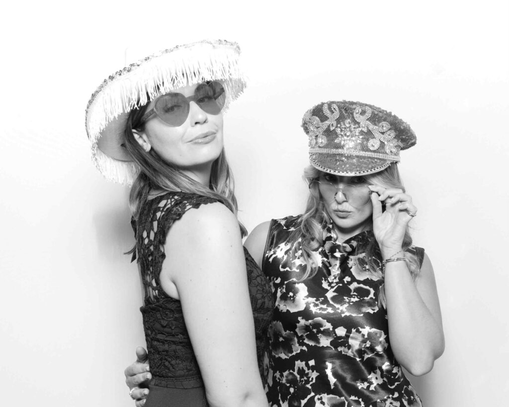  Two women dressed in glamorous outfits, wearing fun hats and sunglasses, posing confidently in a sleek black and white photo booth, showcasing creative corporate event trends.