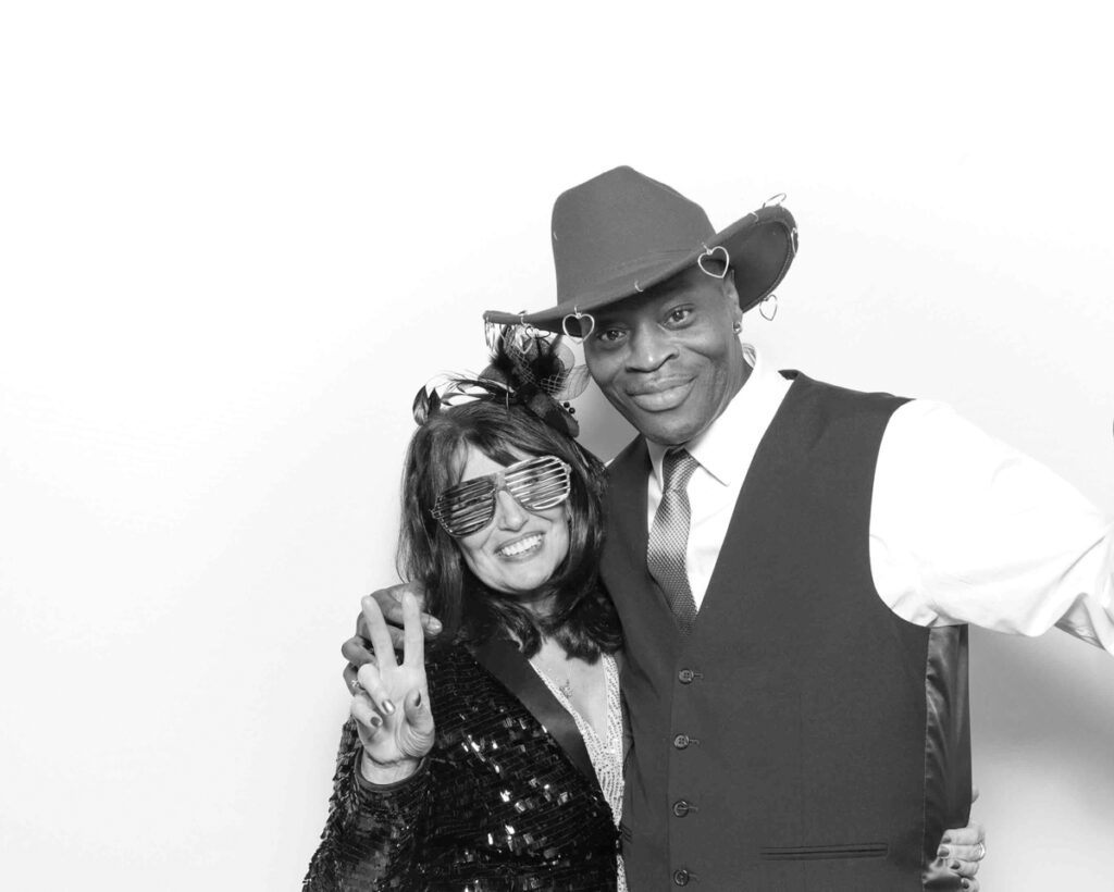  Two guests dressed in playful outfits and fun accessories, striking a pose in a black and white photo booth, showcasing creative ideas for corporate entertainment