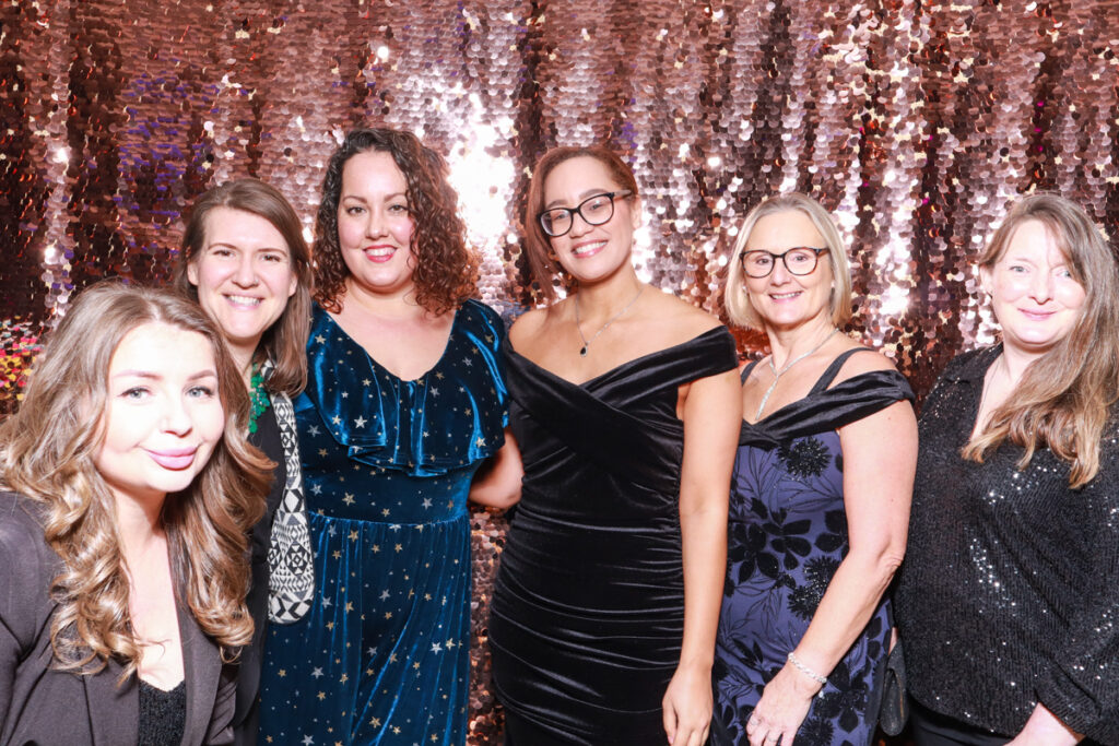 A group of six women in glamorous evening attire posing with a shimmering rose gold sequin backdrop, representing stylish photo booth setups for corporate event trends.