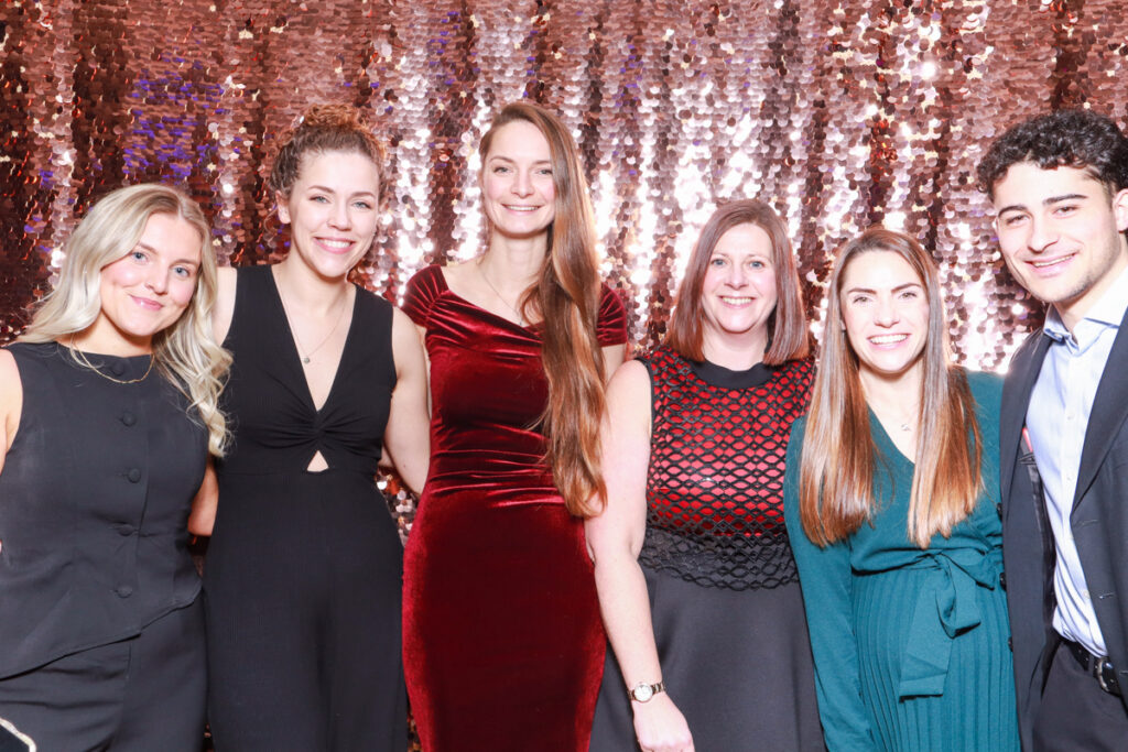 A mix of six colleagues dressed elegantly, smiling in front of a rose gold sequin photo booth backdrop, showcasing fun and sophisticated ideas for corporate event trends.