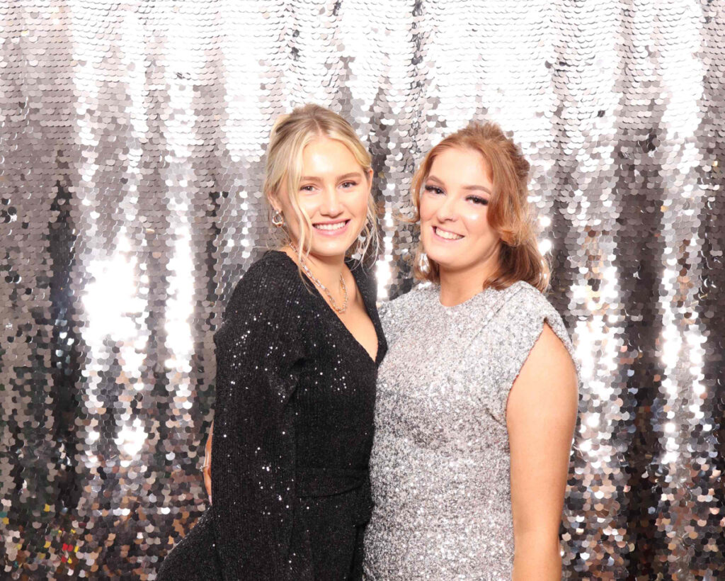 Two women dressed in black and silver sparkling outfits, posing against a shimmering silver sequin backdrop, representing elegant corporate event trends.