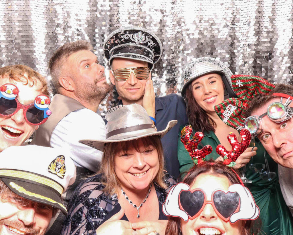 A big group shot with guests wearing festive hats, sunglasses, and props at a Christmas staff party.