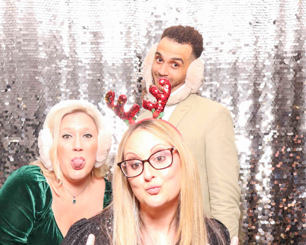 Three colleagues posing with festive props, including reindeer antlers and earmuffs, at a corporate Christmas staff party.