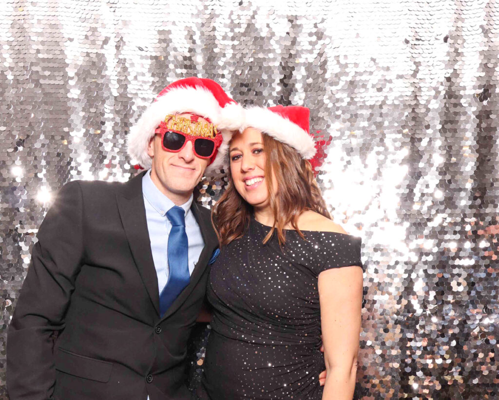 A couple dressed in formal attire, posing with Santa hats and holiday props at a Christmas staff party photo booth