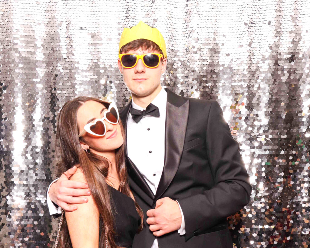 A stylish couple posing with quirky sunglasses and festive crowns at a Christmas staff party.